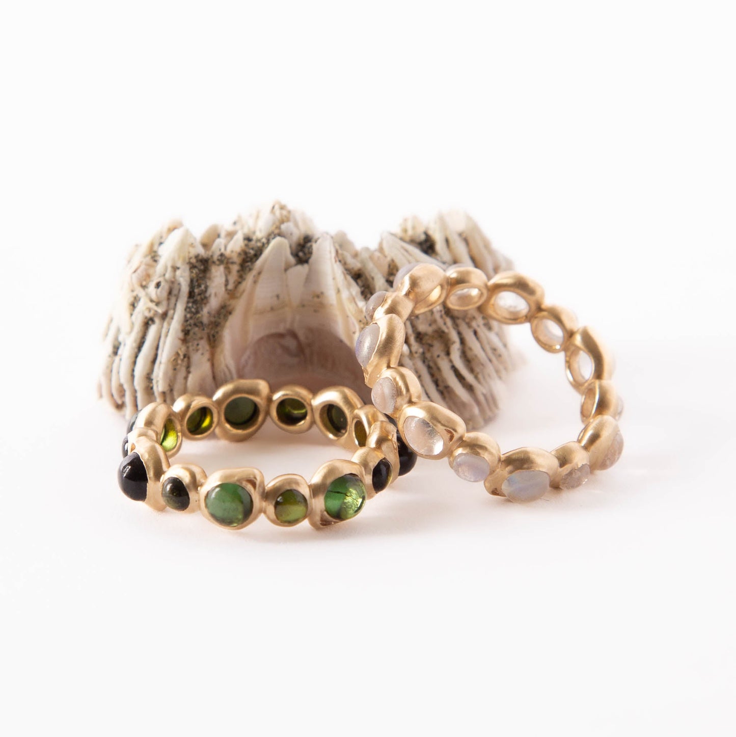 Two gold rings with green and black gemstone accents, displayed on a textured white shell against a white background.