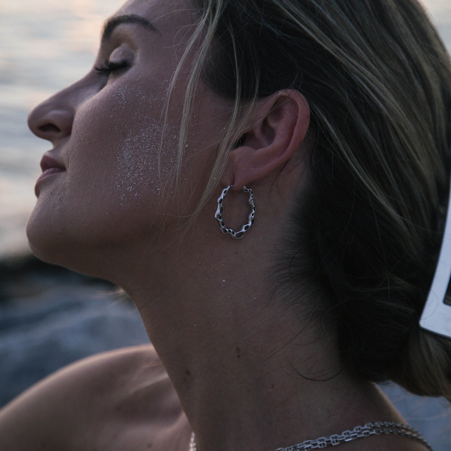 A woman in profile wearing a silver textured hoop earring and a silver chain necklace, with sand on her skin, set against a soft-focus beach or water background at sunset.