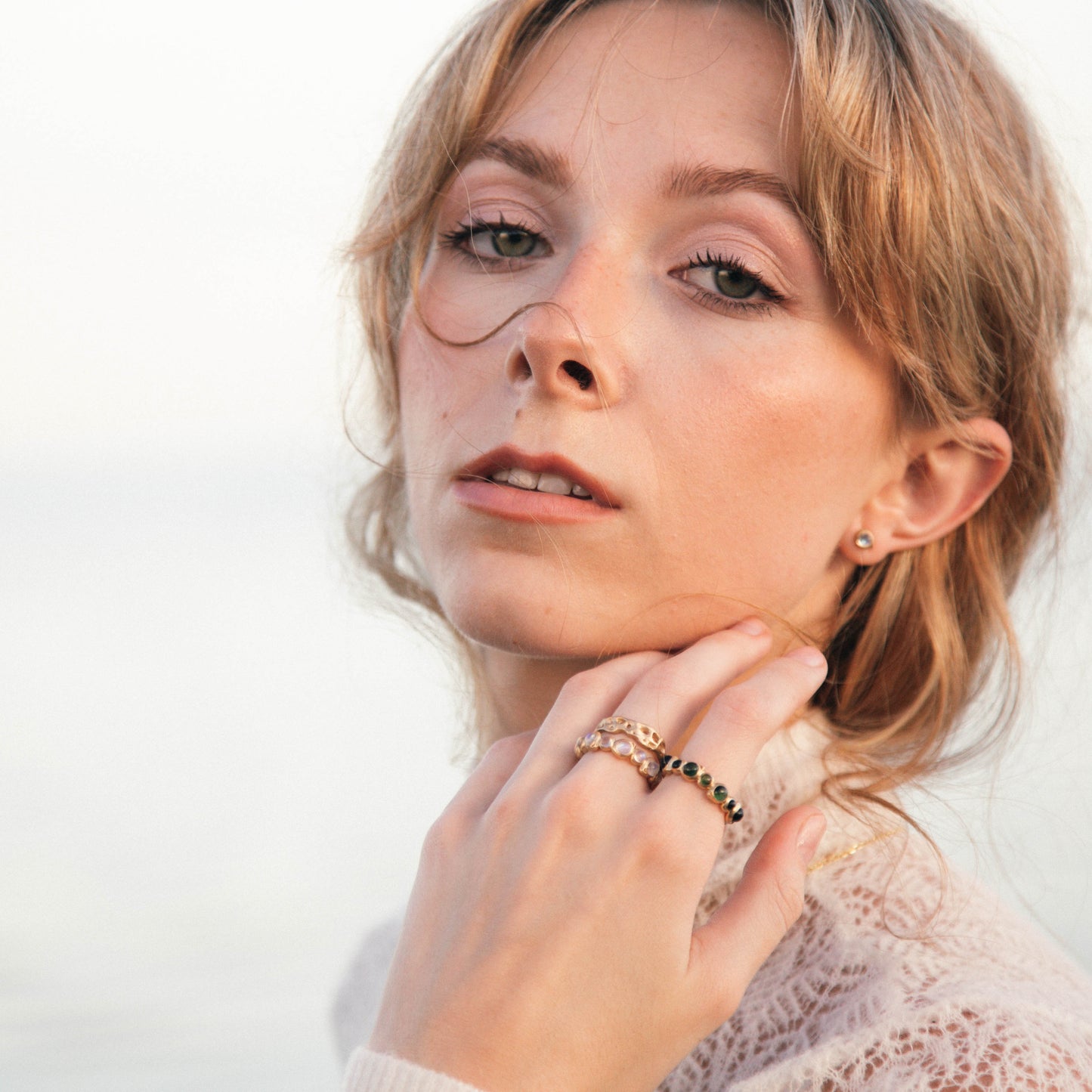 A close-up portrait of a woman with blonde hair wearing multiple delicate rings on her fingers, set against a soft, out-of-focus light background.