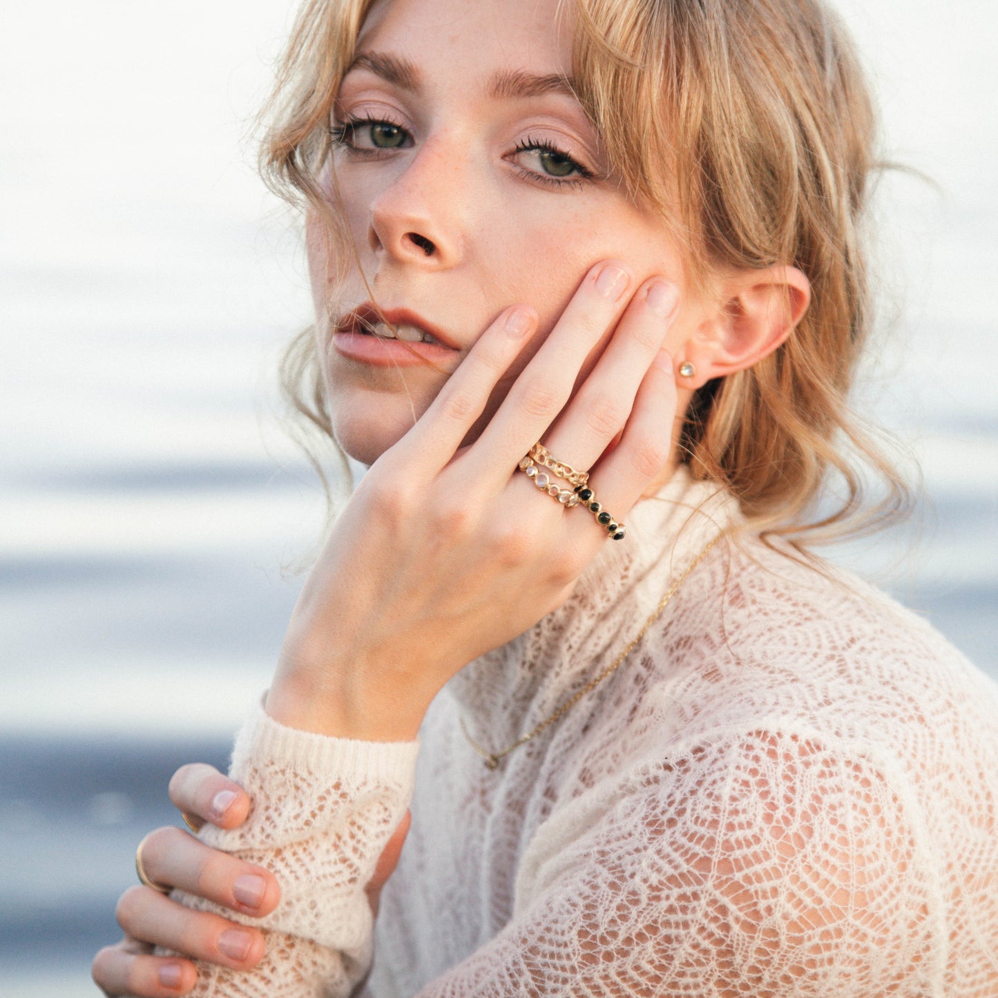 A woman with blonde hair wearing a cream lace-knit sweater and gold rings, posing outdoors with a soft-focus background of water or sky.
