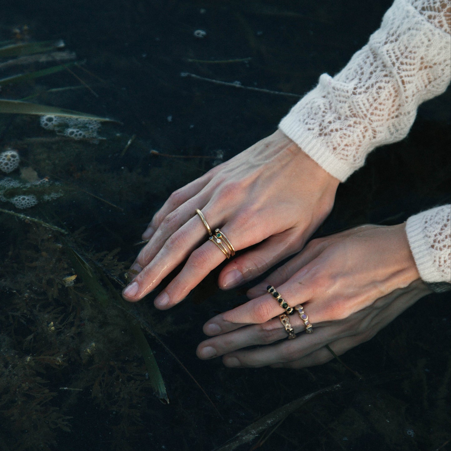 A close-up lifestyle shot of a person's hands resting in dark water, wearing multiple gold rings, including a stack on one finger and a beaded ring on the other, with a white lace sleeve visible.
