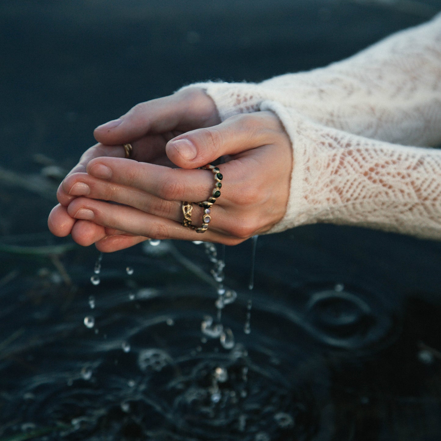 A close-up lifestyle shot of a person's hands cupping water outdoors, wearing a cream-colored lace-knit sweater and several gold rings.